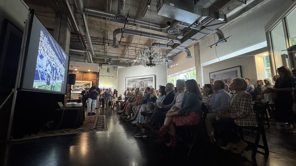 A seated audience facing a large projection screen in a room with an exposed ceiling