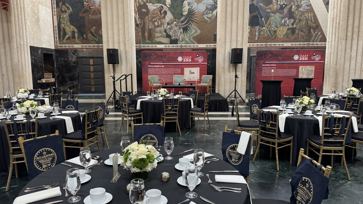Speakers on stands either side of a small stage in a private dining room set with round tables and gold chairs