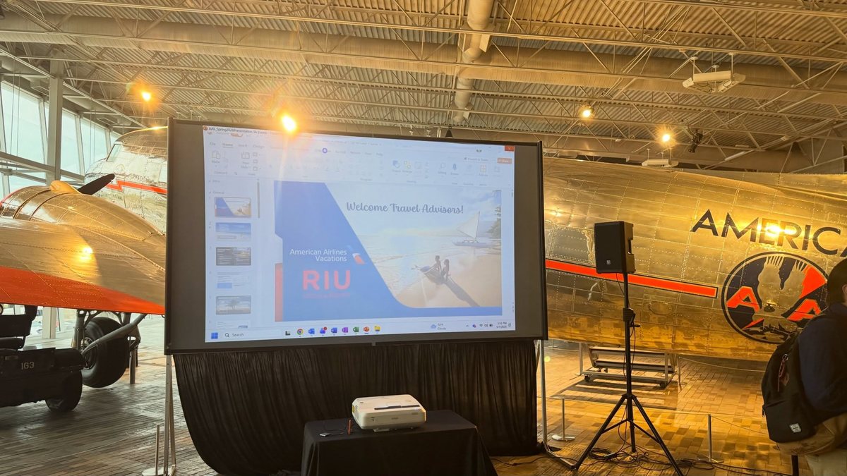 A projection screen and a speaker on a stand set up in front of an aircraft in a hangar