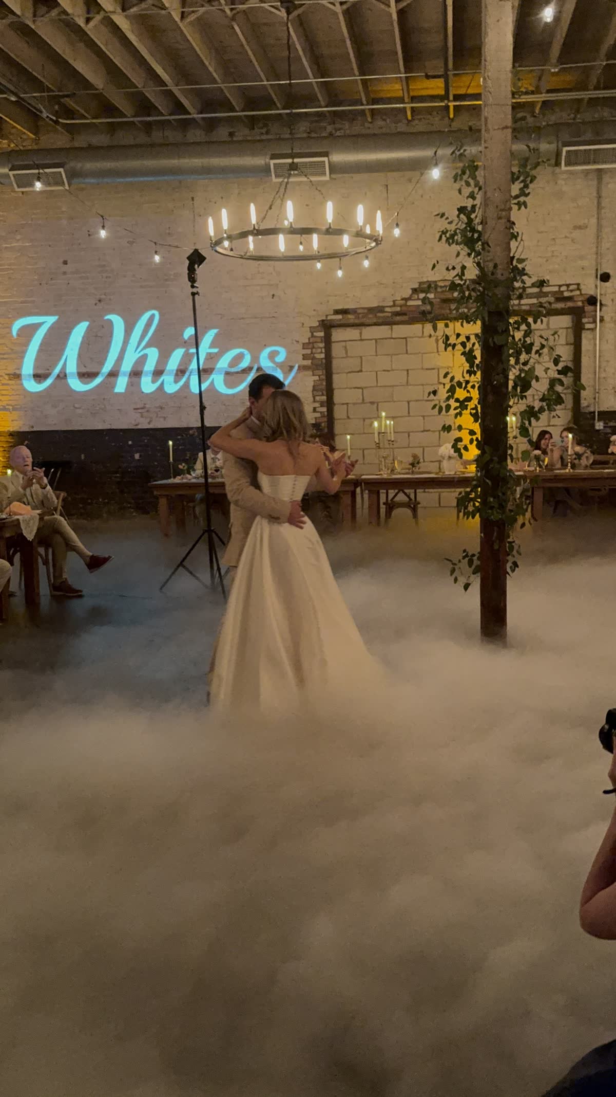 First dance on a low lying fog cloud in an industrial brick loft, with a cyan monogram projected on the wall behind the couple