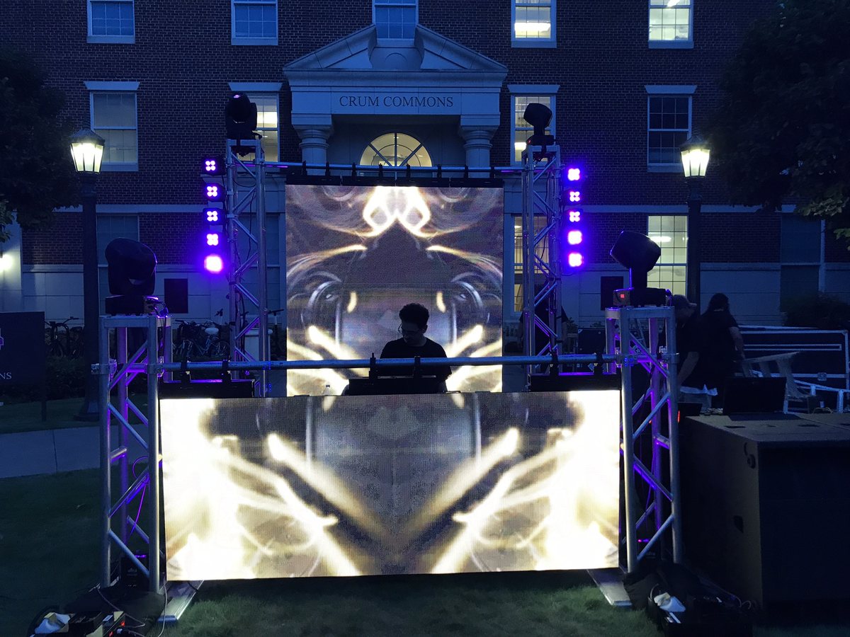 A DJ playing behind a lit booth with an LED wall and truss towers at an outdoor evening event
