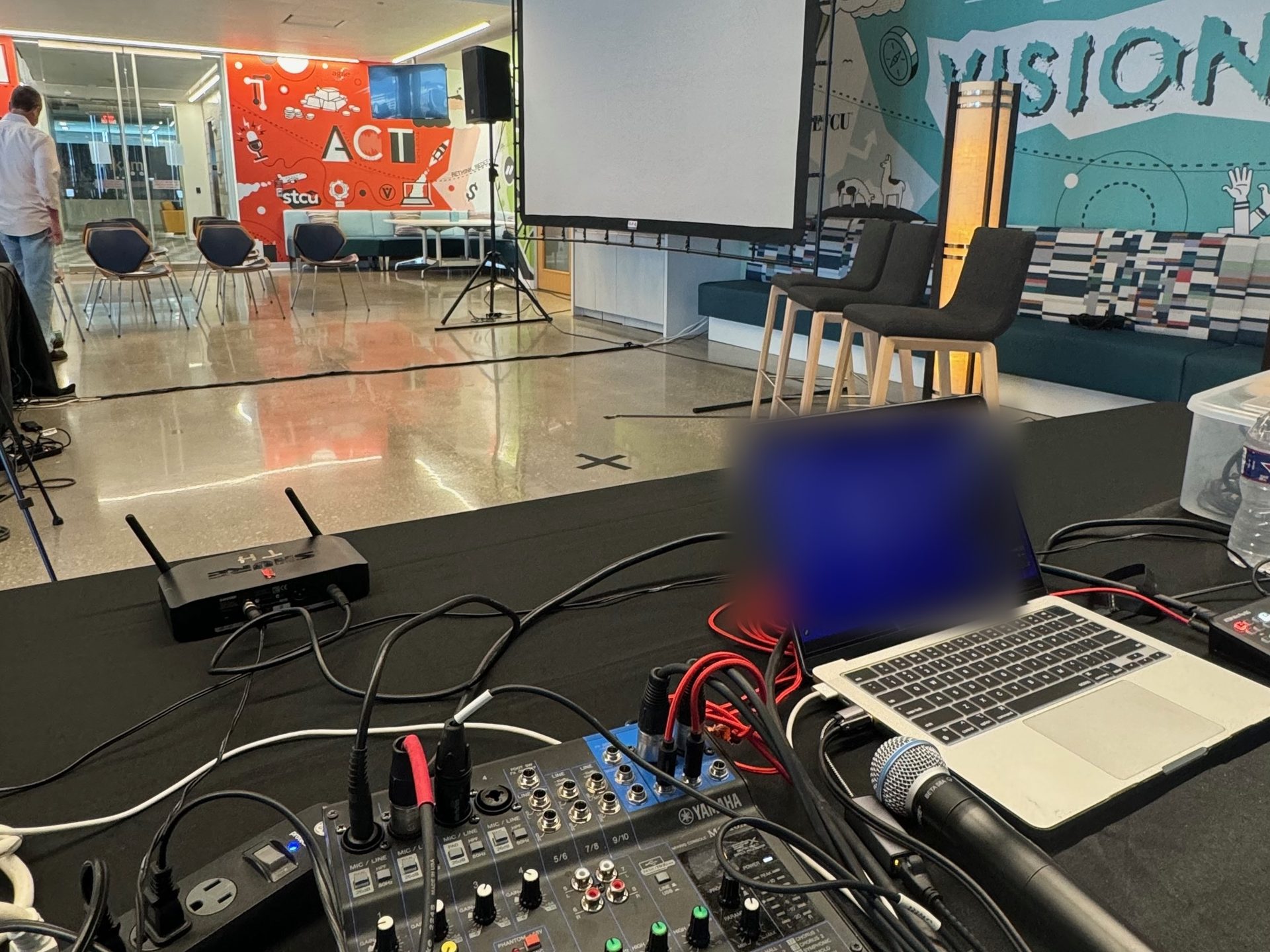 A Yamaha mixer, wireless handheld and streaming laptop in front of a panel stage with a projection screen at a Dallas office event