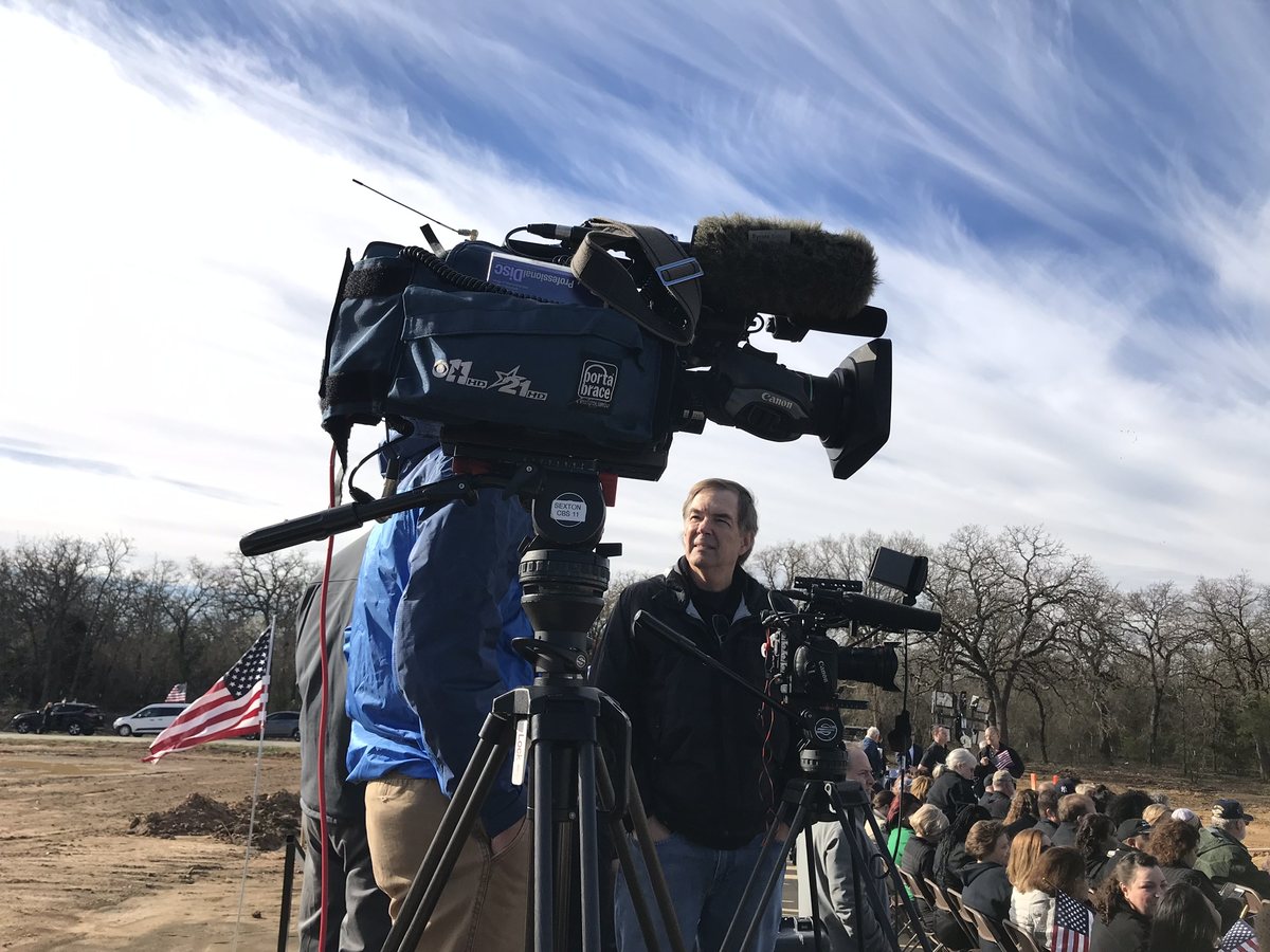 A video camera on a tripod with its operator, covering an outdoor event with a seated audience behind