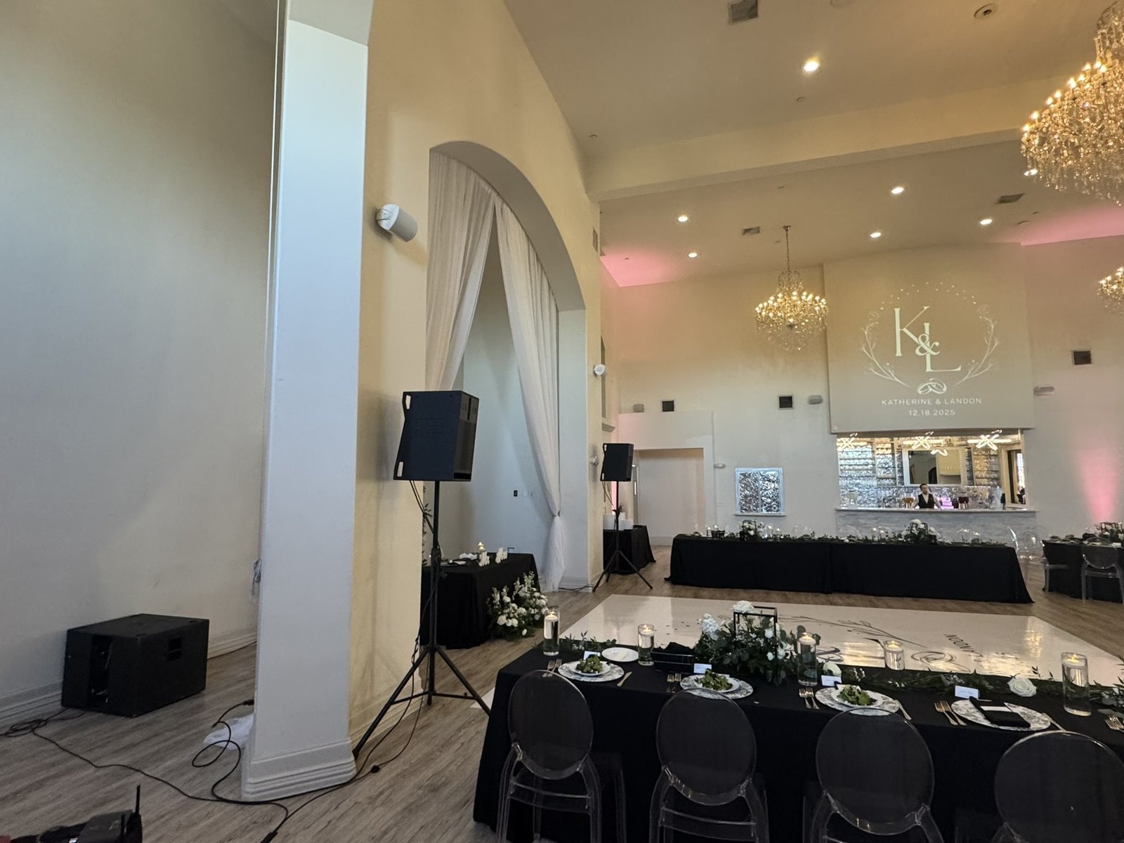 A speaker on a stand and a subwoofer beside a white dance floor, with a monogram projected on the wall behind