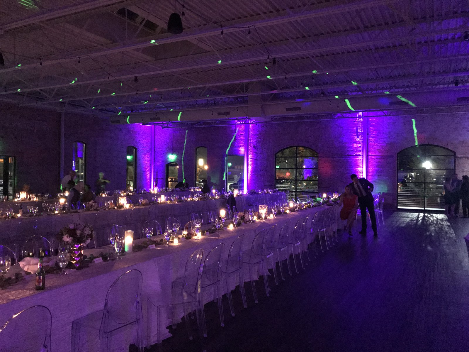 Purple uplighting along a long candlelit banquet table under an open industrial ceiling