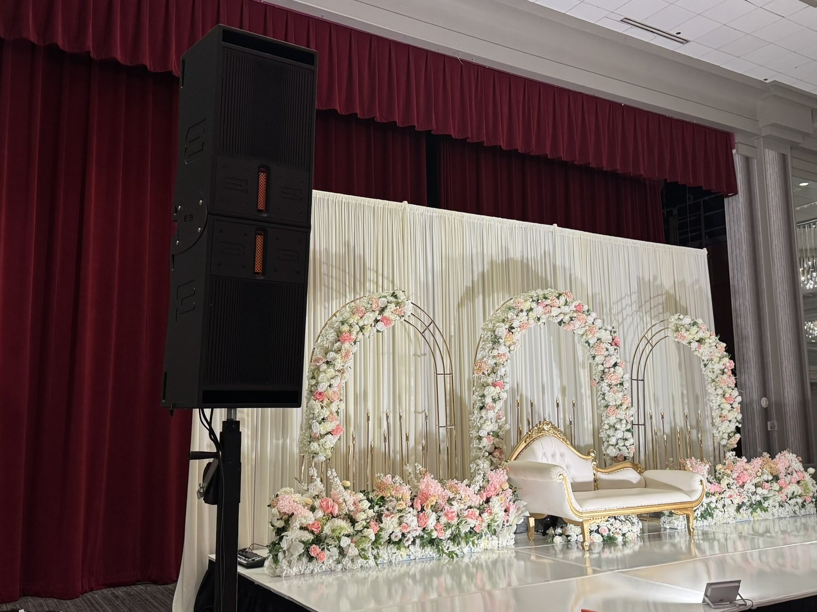 A speaker column beside a floral ceremony arch and a two seat sofa on a white stage