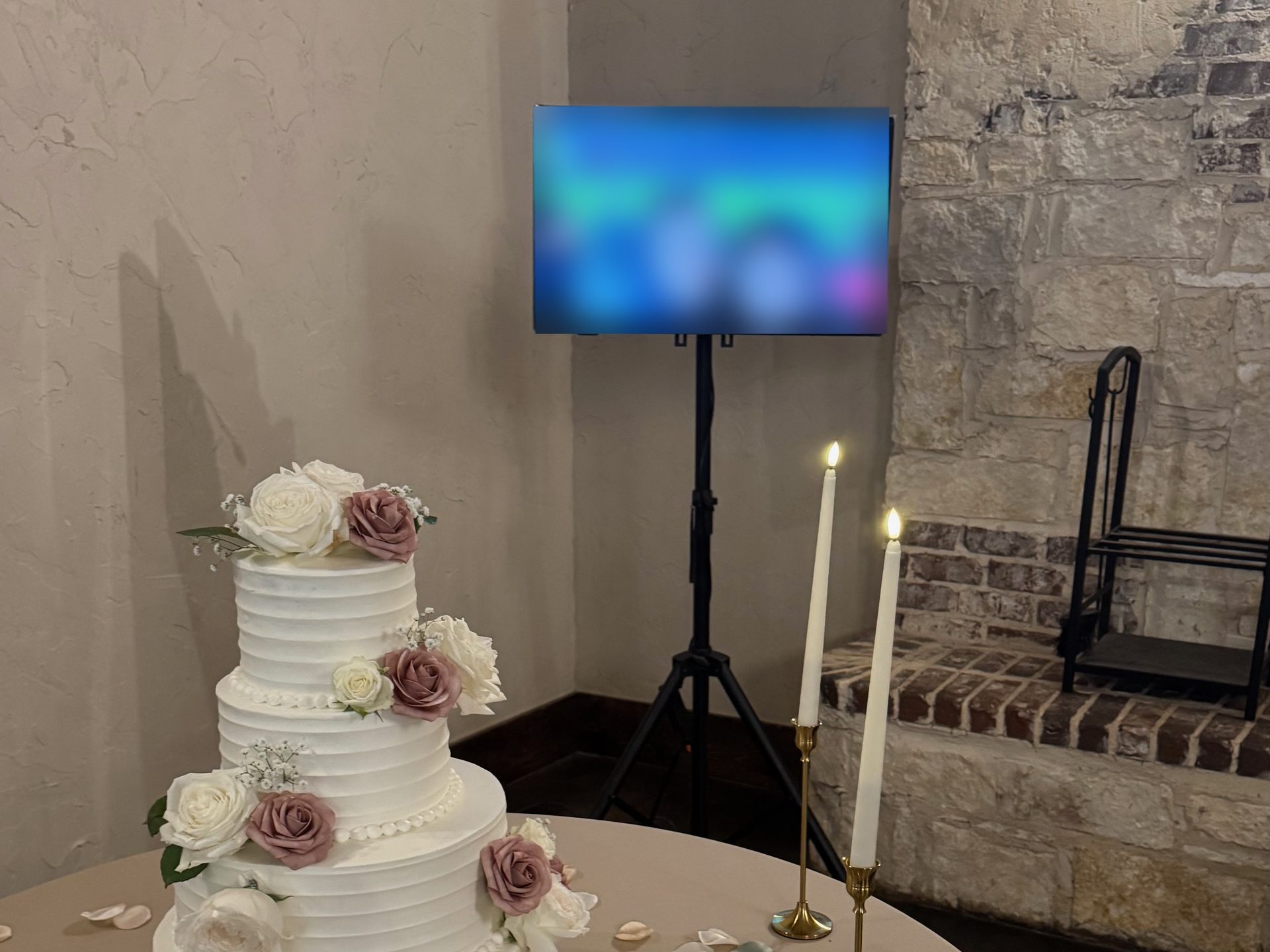 A television on a floor stand playing a photo slideshow behind a three tier wedding cake and taper candles