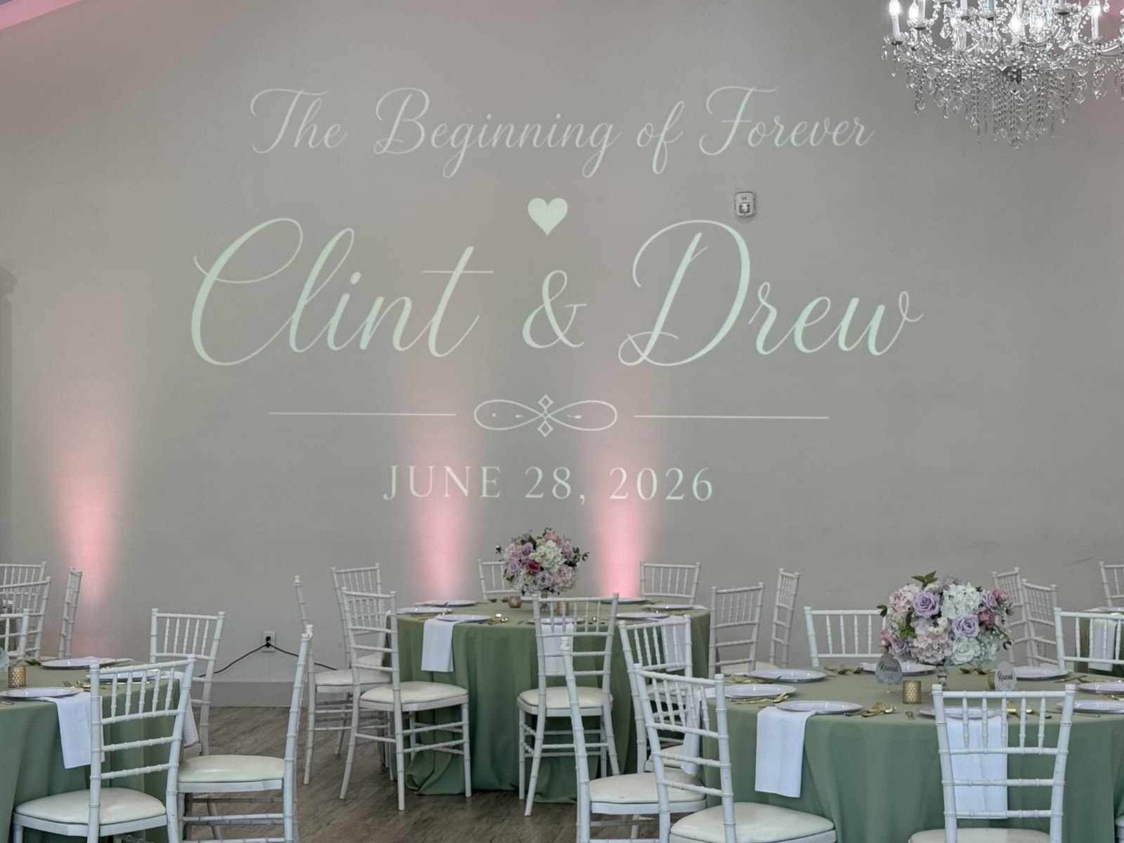 A wedding monogram projected across a reception wall above pink uplights, with tables set in sage and white