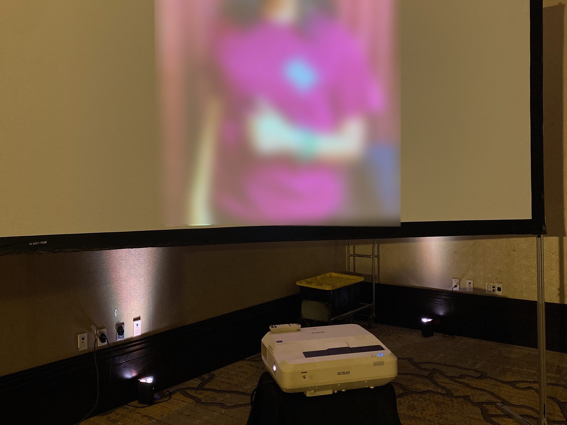 An ultra short throw projector on a skirted table at the base of a fast-fold screen showing a wedding slideshow in a hotel room with uplighting